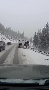 Tire Cracks on Ice — Truck Loses Control The dashcam footage captures a winter drive along a snow-covered mountain highway with steady traffic moving through icy conditions. The road surface is slick, with patches of hard ice and thin layers of snow. Pine trees line both sides of the roadway, and a metal guardrail separates the right lane from a steep, snow-covered slope below. As traffic moves downhill through a gentle curve, a white semi-truck with a long box trailer is traveling among other v
