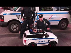 2 NYPD OFFICERS, INTERACTING WITH 2 CHILDREN DRESSED IN UNIFORM RIDING AROUND IN A TOY NYPD CAR.