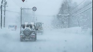 Snow covered vehicles driving cautiously during heavy snowfall through small town. Harsh winter conditions with huge piles of snow on roadsides and low visibility. Challenging car ride in wintertime.