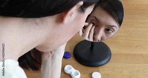 Close Up headshot portrait of a brunette woman taking off her contacts and putting them in the plastic container. Girl's face is reflected in the make-up mirror, shooting from behind