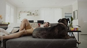 Young adult bearded man hanging out with his Great Dane dog. Hipster male spending quality time with his four legged pet friend, laying on the couch in living room and browsing cell phone