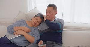 senior retirement Asian married couple resting on a couch, the man comforting the woman who has a headache.