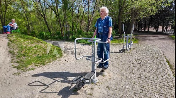 Old gray-headed male senior in his seventies (75) in blue jeans pants and jacket exercising on a hip twister in the outdoor fitness gym in a public park and his wife resting on a bench. 4K video