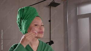 Portrait of happy attractive middle aged woman in bathrobe and towel wrapped around head cleansing skin with cotton pads, making spa treatment and facial skin care in bathroom.