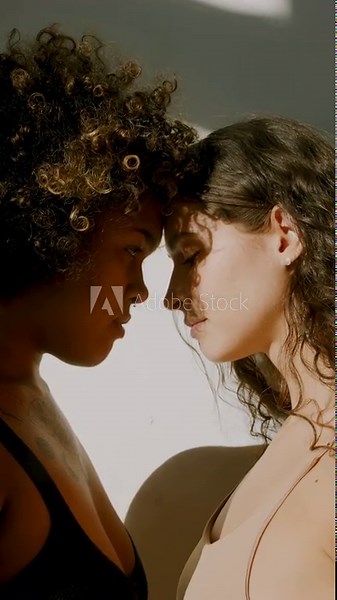 Vertical close-up side view portrait footage of two ethnically diverse young women standing in front of each other head to head opening eyes looking at each other