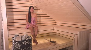 Beautiful Filipina at spa treatment in wooden Finnish sauna wearing pink towel. Attractive young woman sitting on the bench in a hot sauna and relaxing. Healthy weekend at beauty spa centre and sauna.