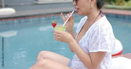Woman in white coverup by pool holding cocktail, camera panning, stirring straw, sipping, cooling