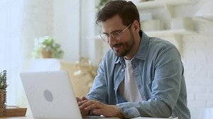 Concentrated young man freelancer in eyeglasses looking at laptop screen, working remotely at home. Head shot focused smiling guy chatting in social network, typing message, internet web surfing.