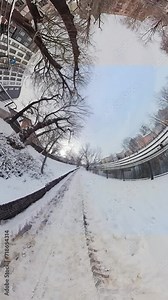 A 360-degree video of a snowy path in a city park. The path is covered in snow and there are trees and buildings on either side. The video is taken from the perspective of a person walking down the