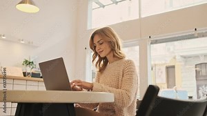 An attractive blonde woman is typing on her silver laptop computer while sitting in the white cozy cafe