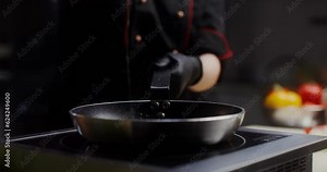 A woman in a chef's uniform with disposable black gloves on her hands pours oil on a frying pan standing on the stove, a close-up of her hands