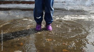 The child jumps from happiness in a puddle. The child jumps merrily through a puddle. Spring childish mood. Boots in a puddle close-up. Splashes in all directions, slow motion.