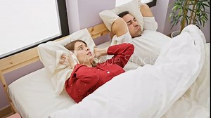 A woman annoyed by snoring lies awake next to a sleeping man in a bedroom setting, depicting a common couple's issue.