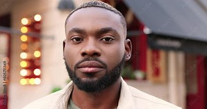 Portrait of serious African American young handsome man looking at camera and standing at street near cafe. Close up of good-looking male outdoor. Person in town outside.