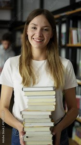 Student girl walks through library with stack of books in hands, shelves with book, front view. Young woman hold books at hands walks through library