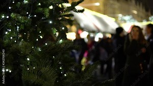 A tree branch decorated with Christmas lights, people walk at a Christmas market in the background.