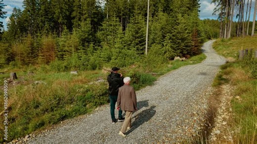 Male walking with senior woman along gravel trail in forest under sunny weather. Elderly lady strolling beside adult son on peaceful woodland path beneath clear sky. Man and woman hiking through tree
