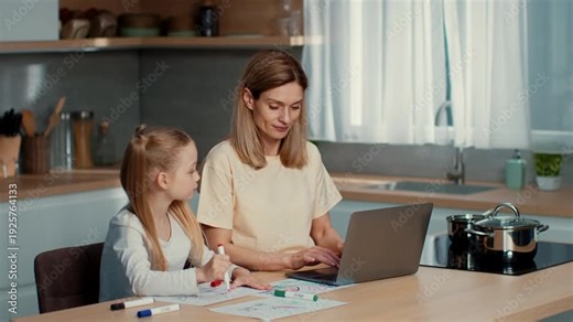 A woman with light skin works on her laptop at a kitchen table while her daughter with long blonde hair draws pictures on paper. The kitchen is warm and homey.