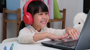 Download Cute elementary school girl wearing headphones and using a laptop computer. Happy Asian kids study online interactively with laptop computer or homeschooling, listening to music or playing games. for free