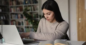 Focused fresh gen Z Latin college student girl writing notes, lesson summary, using laptop in public library, typing, watching webinar, online class, turning face, looking at camera, smiling