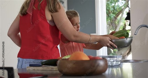 Woman and child girl moving colander rinsing greens under faucet at kitchen sink making meal