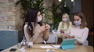 Women in coronavirus face masks disinfecting hands with sanitizer sitting in cafe indoors. Middle Eastern and Caucasian colleagues meeting for planning in restaurant on Covid-19 pandemic lockdown