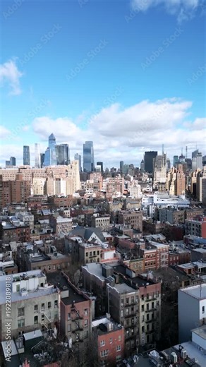Aerial view moving backward over the historic rooftops of greenwich village, revealing the expansive and iconic midtown manhattan skyline under a bright, partly cloudy blue sky in new york city