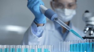 Female scientist researcher wearing a lab coat, mask, and safety glasses is using a micropipette to drop a blue liquid into a test tube, close up view. Medicine and science concept
