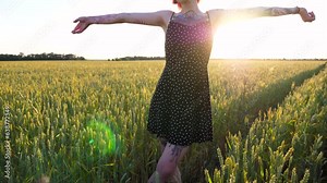 Pretty hippie girl in dress standing on green barley field and raising hands at sunset. Happy punk woman with tattoos enjoying freedom on wheat meadow at summer. Scenic rural landscape at background