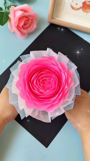 Let’s create a beautiful rose bouquet! Start by taking 40 sheets of 5 by 5 cm paper. Cut the corners of each sheet to give them a more rounded shape. Next, cut the bottom off a cup to use as the base, and start placing the sheets on it one by one, forming the petals. Use double-sided tape to secure each layer before adding another, continuing this process until your bouquet takes on the desired fullness. Once the base is complete, cut the top off a plastic bottle to create a sturdy stem, and att