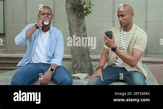 Diverse male friends sitting on bench holding to-go java cups, chatting, glancing at black bag. Urban, plaza, sidewalk, coffee, mug, conversation, casual Stock Video Footage - Alamy