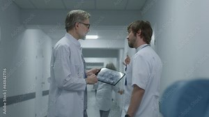 Male doctor shows MRI, CT brain scan image to colleague physician using digital tablet. Medics talk, stand in modern clinic corridor. Medical staff and patients in hospital or medical center hallway.