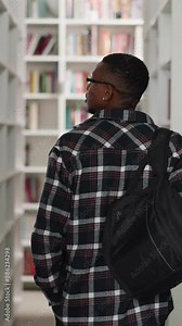 Student walks along aisle in public library. African American man with backpack chooses books on racks in storage. Education and leisure