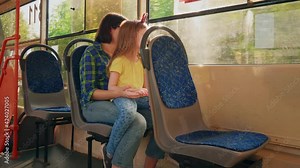 Side view of brunette woman in blue shirt holds her daughter on her lap in streetcar. Family rides in public transport, mom with little blonde girl sit together and look out window tram.