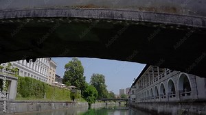 Navigating on Ljubljanica river under famous Triple Bridge. Cityscape view from tourist boat, Ljubljana, Slovenia in hot summer day. Unique perspective of medieval town. Forward moving, slow motion