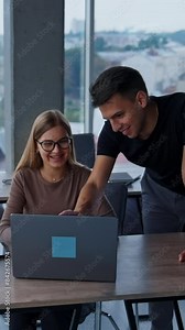 Male colleague bent over the female coworker sitting at desk. People working together discussing something looking at laptop and smiling. Vertical video Stock Video