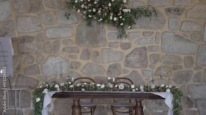 Romantic wedding sweetheart table with floral arrangements, set against a rustic stone wall.