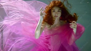 Beauty young woman swimming underwater like in a fairy tale. Underwater view of happy diving girl with long hair in long graceful fuchsia color dress. Slow motion video