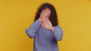 Shocked young adult woman closing eyes with hand showing stop gesture, confused shy scared to watch