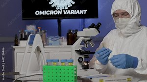 A graphic of the COVID19 mutation or variant named omicron in motion on a monitor in the background of this laboratory where a scientist in full PPE is documenting his research.