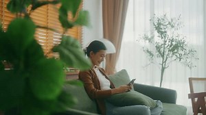 Smiling pretty young Asian woman sitting on cozy couch, using funny mobile apps in living room. Woman at home, Doing online shopping, Messaging friends, Posting on social Media