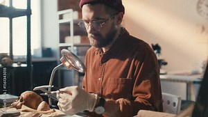 Male archaeologist in gloves and glasses measuring ancient artifact with calipers under magnifying lamp, performing detailed examination at desk in laboratory