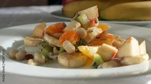 Falling Fruit Salad Ingredients Pieces of Kiwi Orange Banana Apple Seeds Raisins Peanuts on White Dish Plate on Table in Kitchen Dining Room. Vegan Vegetarian Healthy Food. 2x Slow motion 60 fps 4K
