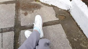 FPV view of the legs in white sneakers, a woman carelessly crossed her legs. Sitting relaxing in the park outdoors on the background of paving slabs.