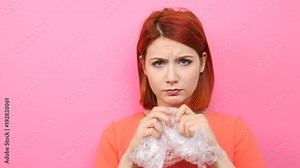 Redhead woman pops a bubble wrap on pink background to calm herself