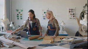 Full shot of two Caucasian female leather workshop employees standing at cutting table with laptop, looking at pieces of leather and suede, and discussing new product designs