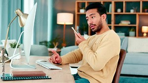 Young IT specialist talking on a phone call while working on a desktop computer alone at home. One young Asian computer programer having a conversation and typing an email while working a desk