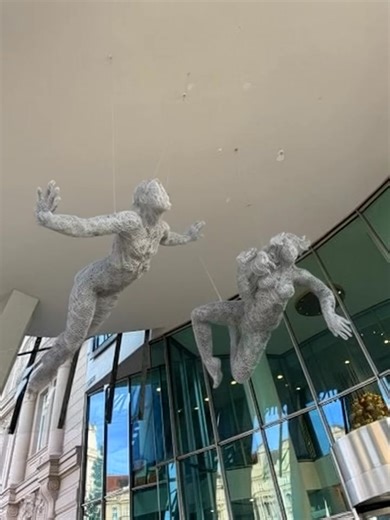 EN: Two light figures float beneath the “skirt” of the Dancing House and suspended between the sky and the Vltava River, where architecture meets the wind. The air currents gently move them, making their bodies appear as if they are truly swimming through space. “The Swimmers” reflect both freedom and solitude both together, yet each alone, carried by an invisible current. The installation responds to a unique site where light, wind, and architecture shape a sculptural narrative. Veronika Psotko