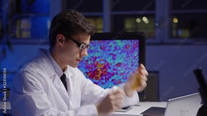 Tilt up side view of young scientist in lab coat and eyeglasses examining test tube with yellow liquid, typing on laptop and working on computer with magnified microorganisms on screen in laboratory