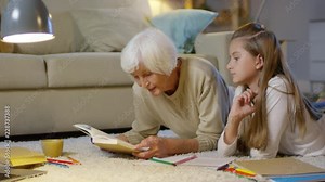 Happy grandmother lying on the floor in cozy living room and reading textbook to granddaughter while doing homework together